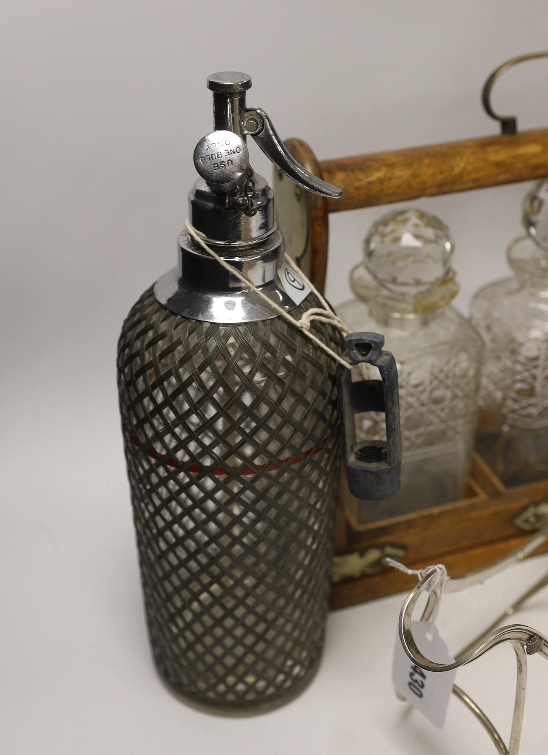 An oak three bottle tantalus, a German 800 standard white metal bottle cage and a vintage soda syphon, 36cm wide